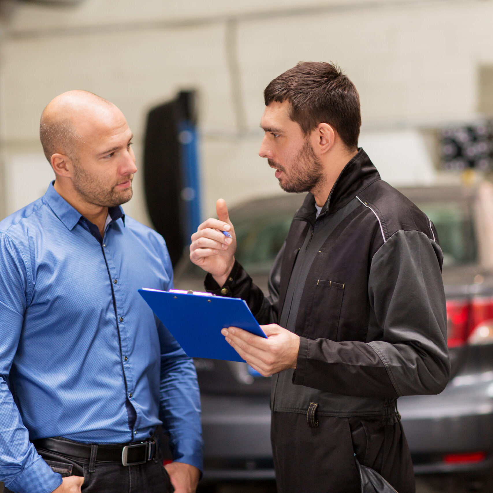 auto service, repair, maintenance and people concept - mechanic with clipboard and customer or car owner at workshop
